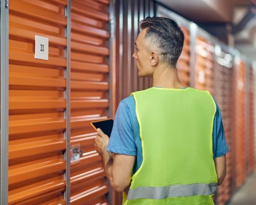 Warehouse security. Man in bright vest with tablet working in warehouse guard standing with back to camera near garage door